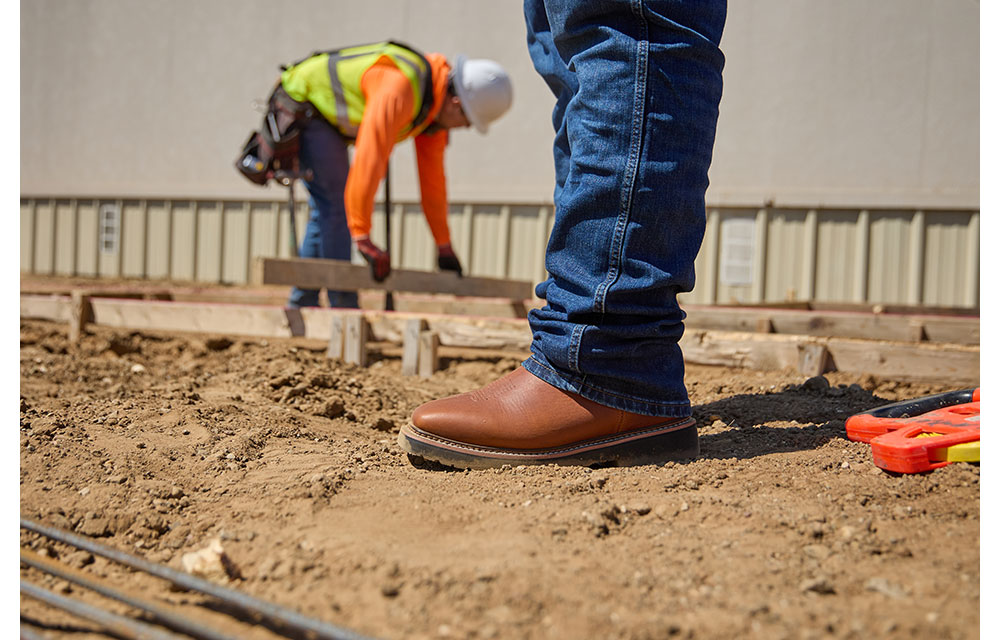 Close-up of Benny Aguilar wearing brown leather Homeland work boots and jeans standing on a dirt construction site.